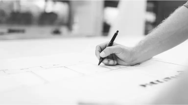 Close-up of a professional’s hand holding a black pen and drawing on architectural blueprints. The focus on the precise hand movement highlights the meticulous planning involved in structural engineering projects