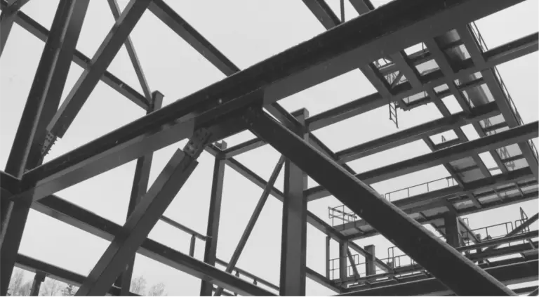 Black and white photograph showcasing the intricate framework of a steel structure under construction. The interlocking grid of beams and columns highlights the complexity and precision involved in structural engineering, demonstrating the skeletal phase before the addition of exterior materials